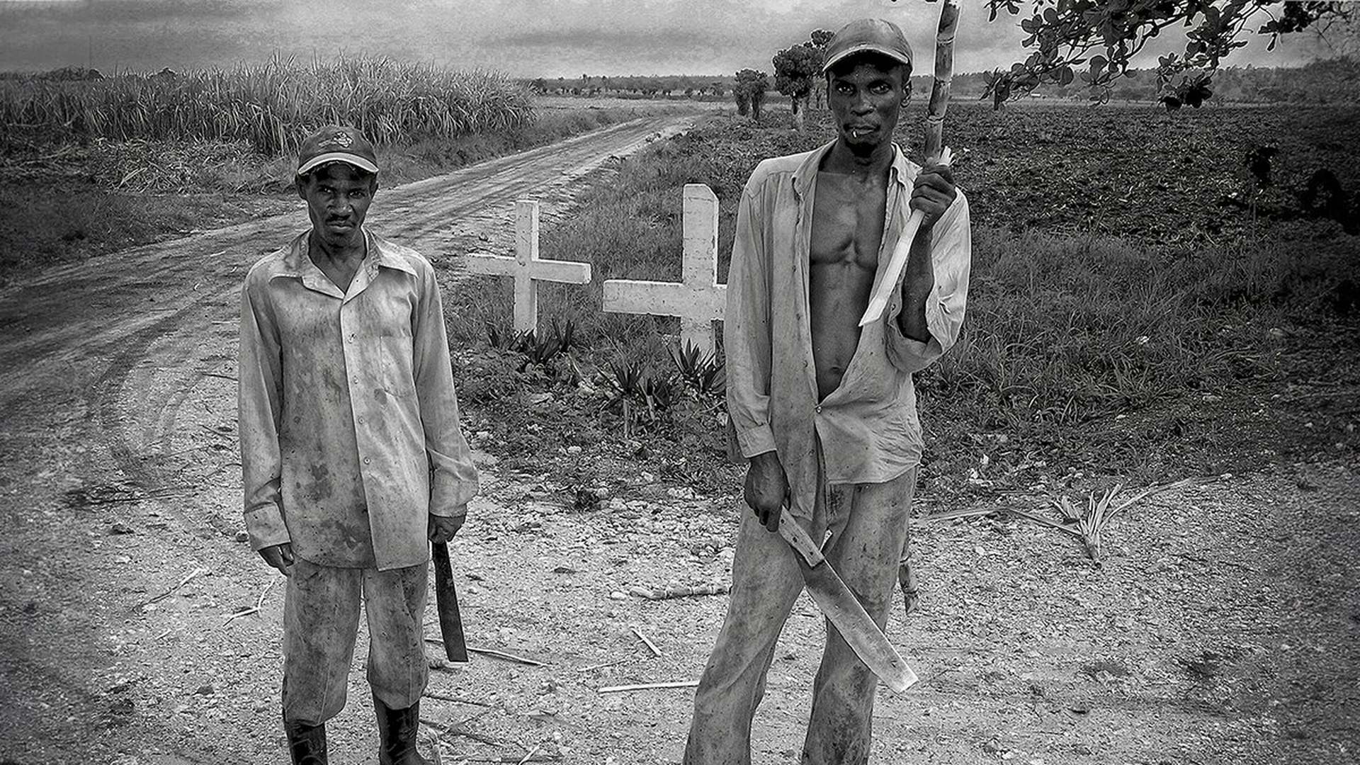 Due tagliatori di canna da zucchero col machete, in piedi sulla strada. Da più vicino avresti solo i volti. Da qui restano dentro anche le due croci.