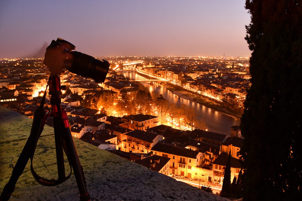 Una reflex sul cavalletto affacciata su Verona, col cielo ancora rosa: sotto, l'Adige, i tetti e le luci della città già accese.