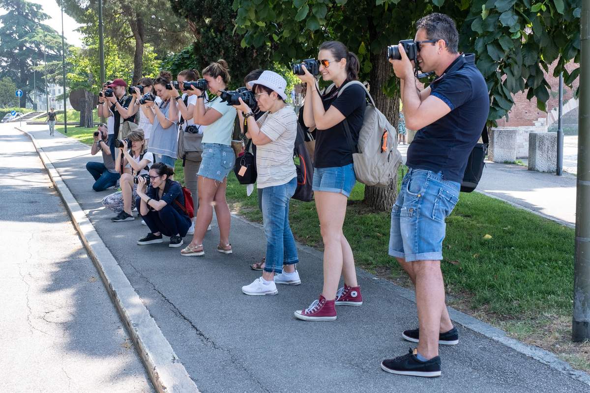 Una fila di corsisti sul marciapiede, all'ombra degli alberi, tutti con la macchina all'occhio puntata dalla stessa parte: una delle uscite.