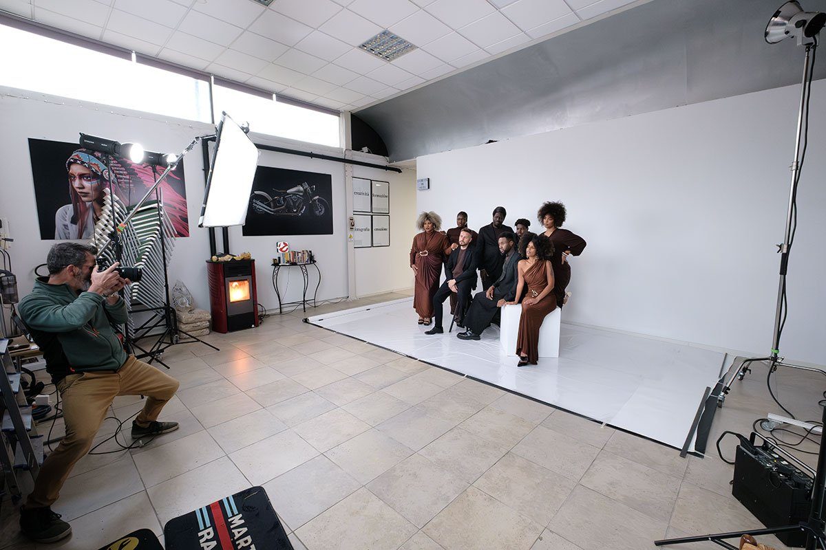 Un fotografo accovacciato con la macchina all'occhio; davanti al fondo bianco, un gruppo di persone in abito elegante sotto le luci già montate.