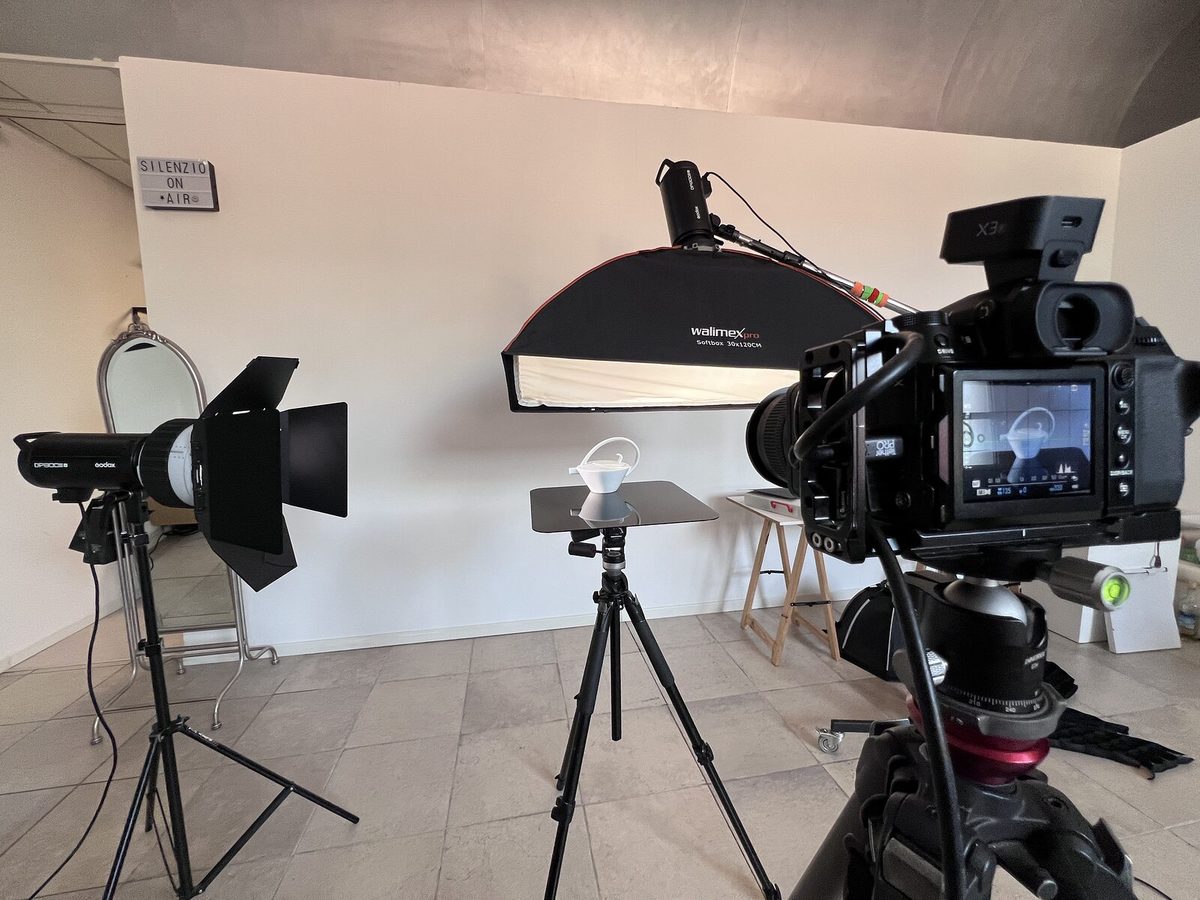 Un set di still life: fotocamera sul cavalletto davanti all'oggetto bianco, bank a striscia sopra e flash con le alette nere sulla sinistra.
