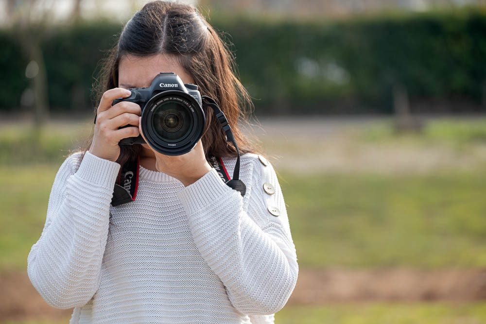 Una ragazza col maglione bianco inquadra tenendo la reflex con due mani, la macchina davanti al viso. Dietro di lei una siepe e un prato.
