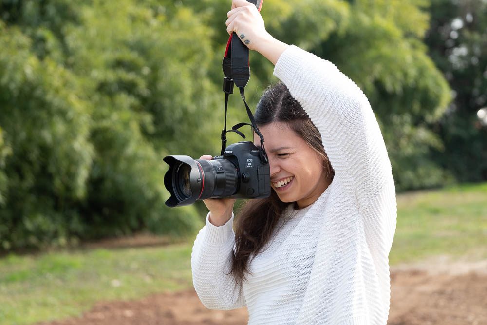 La ragazza col maglione bianco ride mentre tiene la reflex davanti al viso, con un braccio alzato che regge la cinghia. Dietro, alberi verdi e un sentiero di terra.
