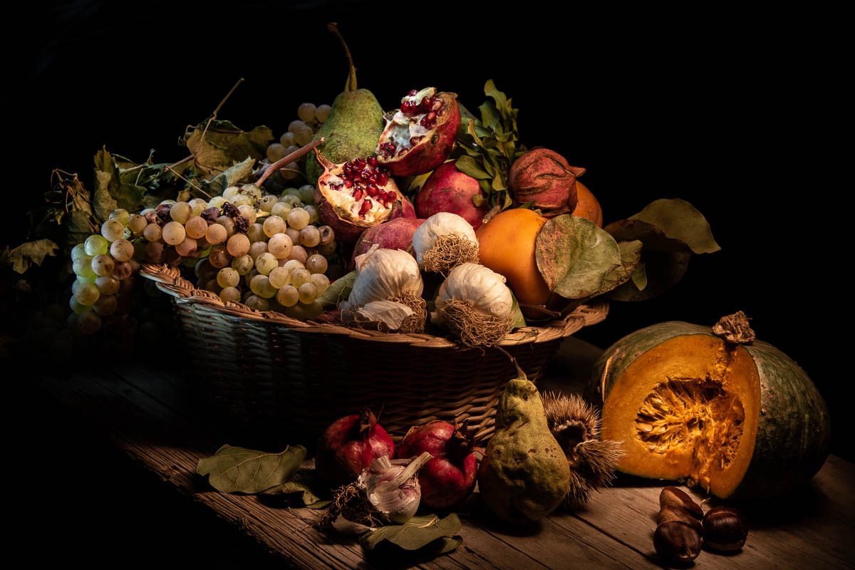 Un cesto di uva bianca, pere, melagrane aperte e teste d'aglio su un tavolo di legno. Accanto, mezza zucca e qualche castagna, e intorno il nero.
