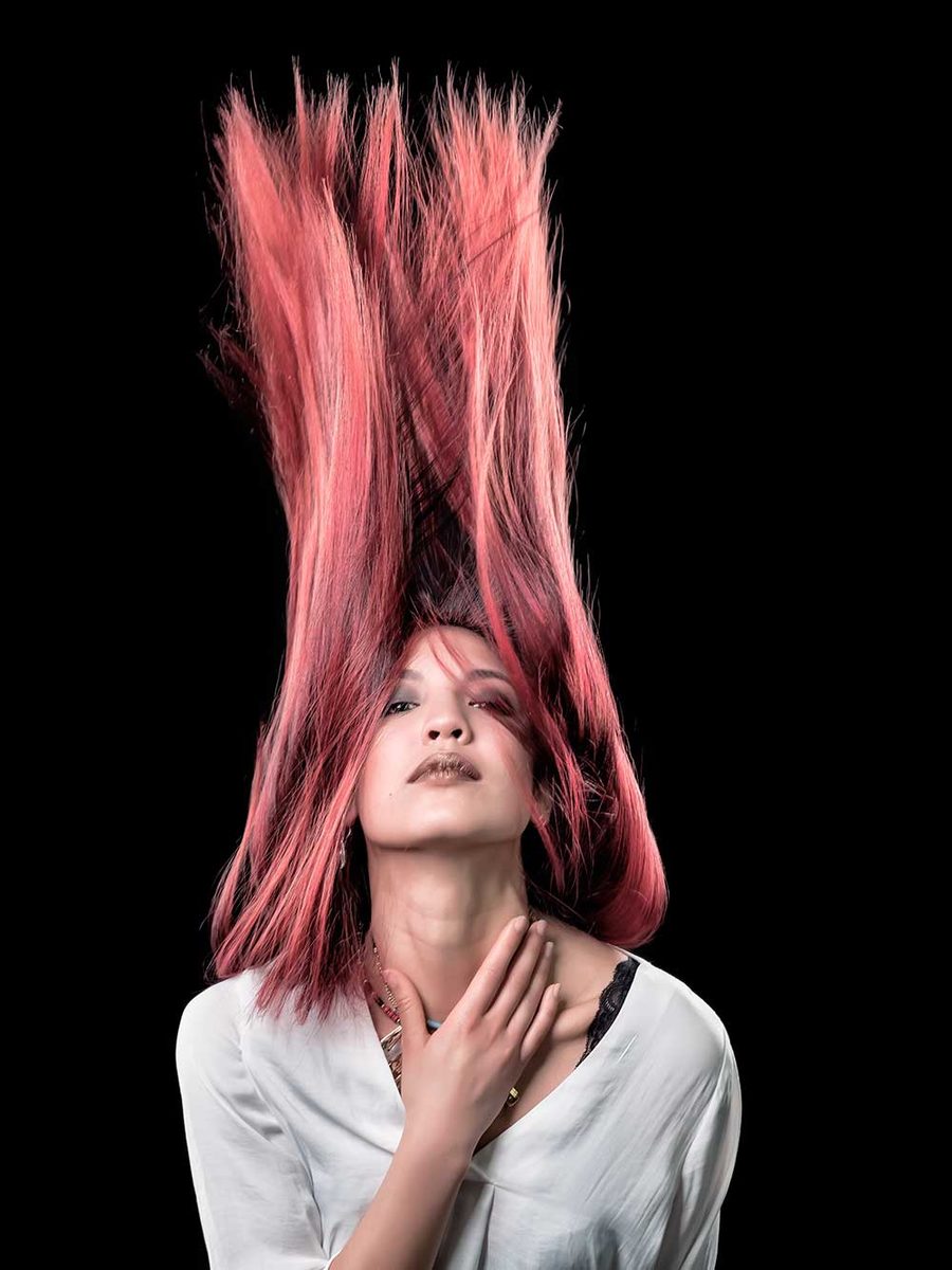 Ritratto in studio di una donna a occhi chiusi su fondo nero. I capelli rosa sono sollevati sopra la testa e una mano è alla base del collo.