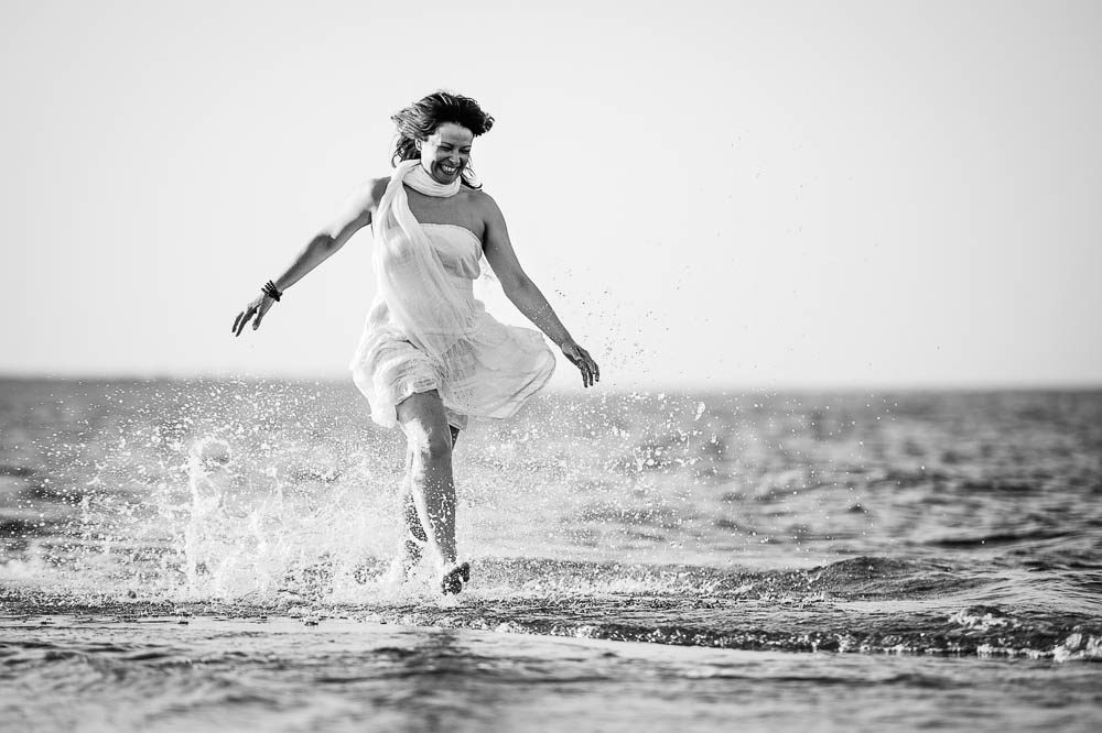 Una donna in abito bianco corre nell'acqua bassa del mare con le braccia aperte. La fotografia è in bianco e nero e gli schizzi circondano le gambe.
