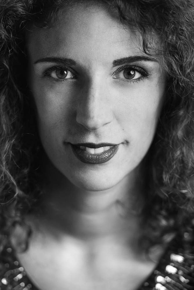 Primo piano in bianco e nero di una donna con capelli ricci e rossetto scuro. Guarda in macchina e i capelli sono illuminati da dietro.