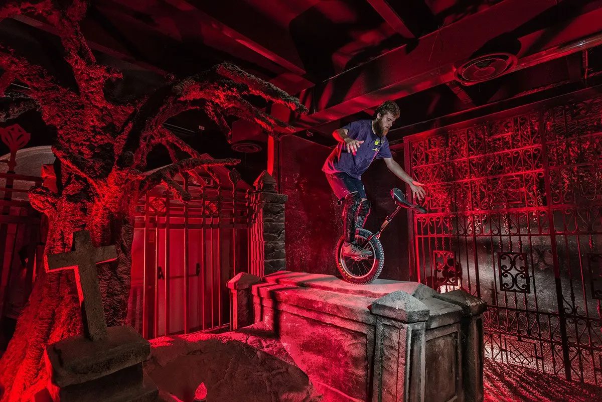 Una scenografia da cimitero annegata nel rosso, e un uomo in equilibrio su un monociclo. Addosso a lui una luce diversa: la maglietta resta blu.