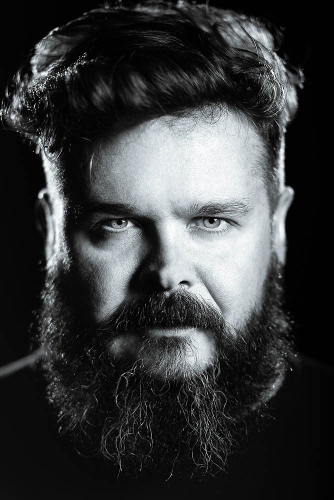 Ritratto in bianco e nero di un uomo con lunga barba e capelli mossi su fondo nero. La luce disegna i bordi del viso e gli occhi chiari guardano in macchina.
