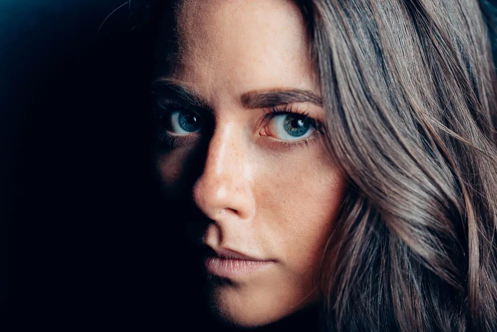 Primo piano di una donna con lunghi capelli ondulati e occhi azzurri su fondo nero. Guarda in macchina con una metà del viso illuminata e l'altra in ombra.