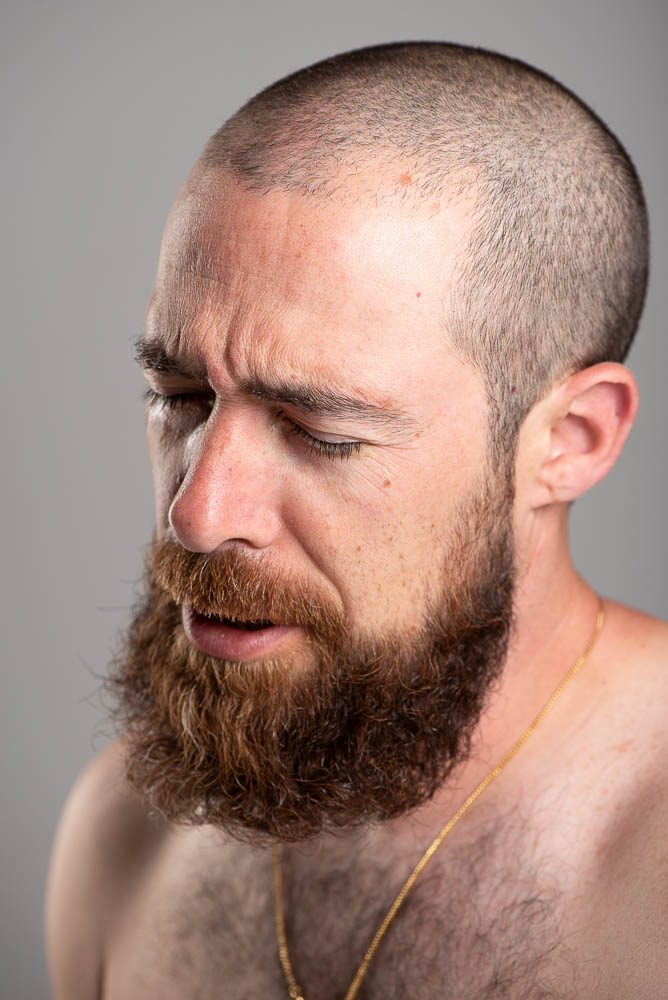 Uomo a torso nudo con testa rasata e folta barba rossa su fondo grigio. Ha gli occhi chiusi e il viso girato verso il basso, illuminato da un lato.