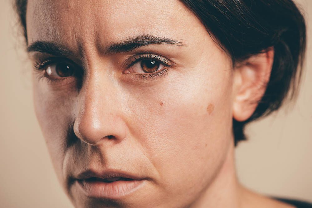 Primo piano stretto di una donna con capelli scuri raccolti su fondo beige. Guarda in macchina senza sorridere, con la fronte leggermente corrugata.